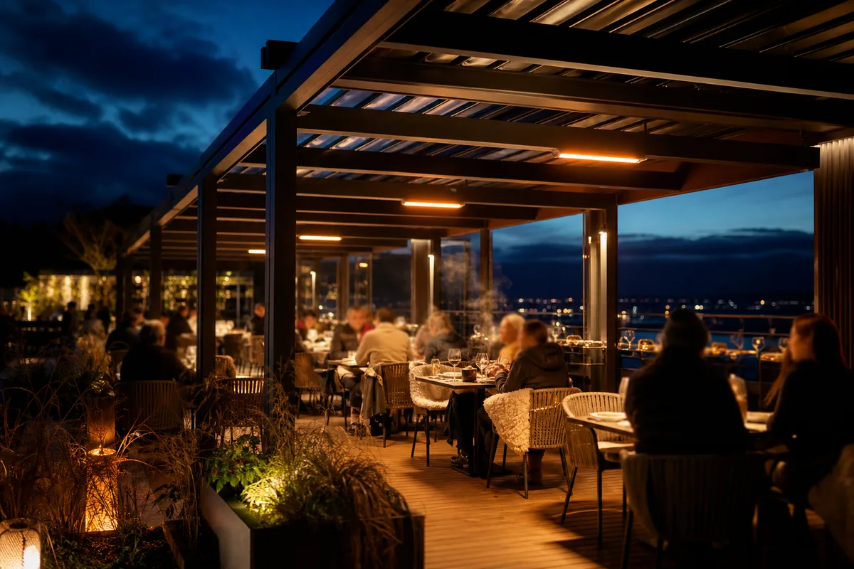 Terrasse de restaurant animée en soirée fraîche, convives attablés de dos sous un éclairage chaud et des radiants électriques discrets